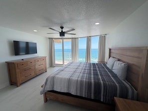 A bedroom with a bed, a wooden dresser, a ceiling fan, a TV, and a balcony with a view of the ocean. - Shores Club 1004 (Daytona Beach Shores)