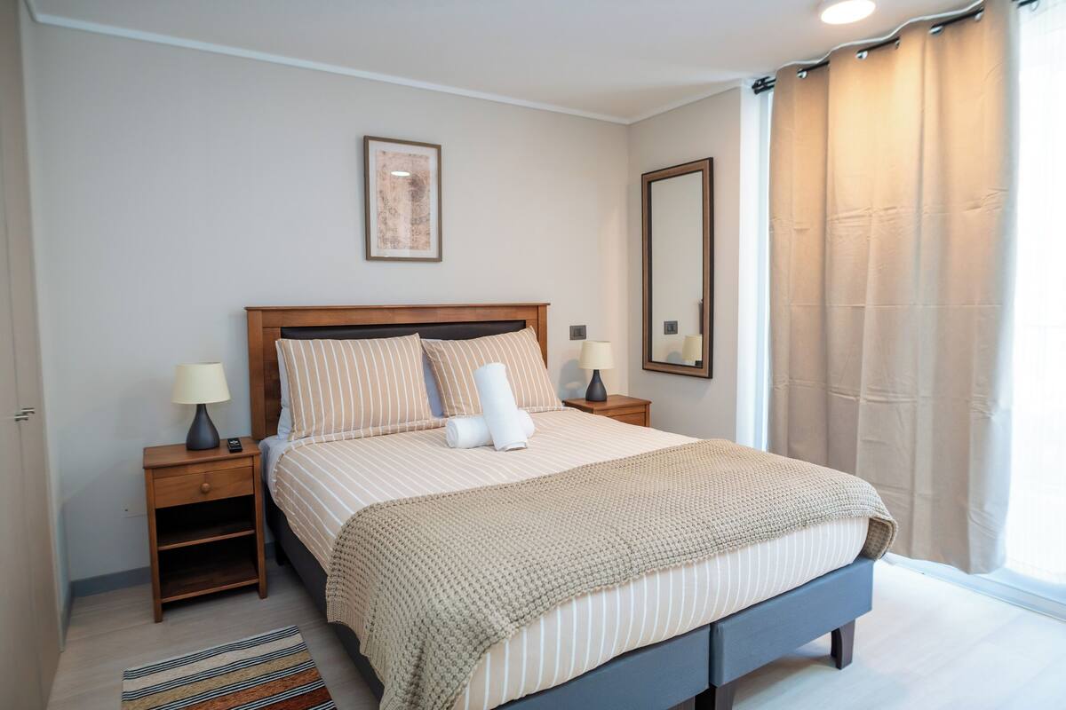 Un dormitorio ordenado con cama, mesitas de noche, un espejo y una ventana con cortinas.