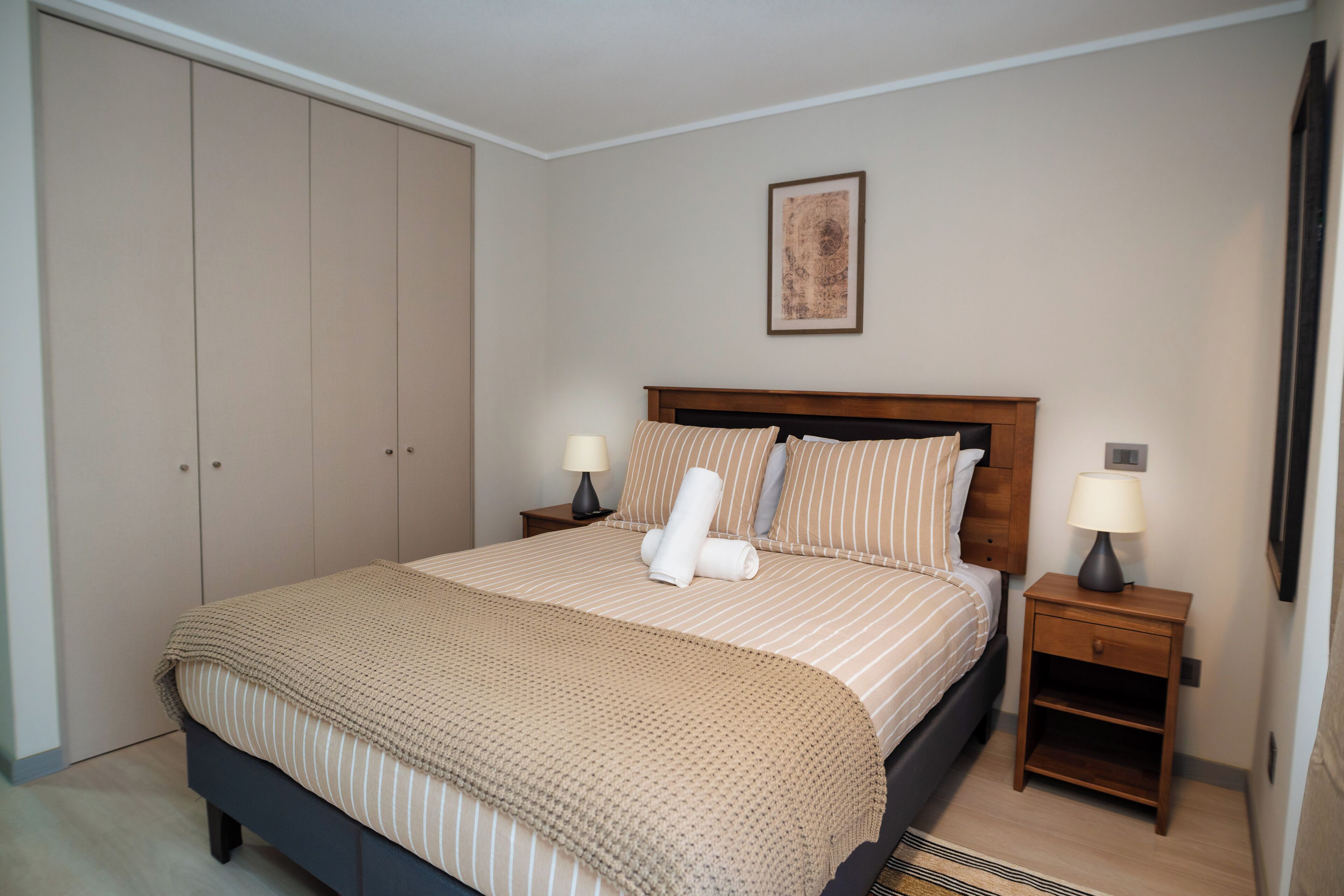 Un dormitorio ordenado con cama, dos mesitas de noche, un armario y un cuadro en la pared.