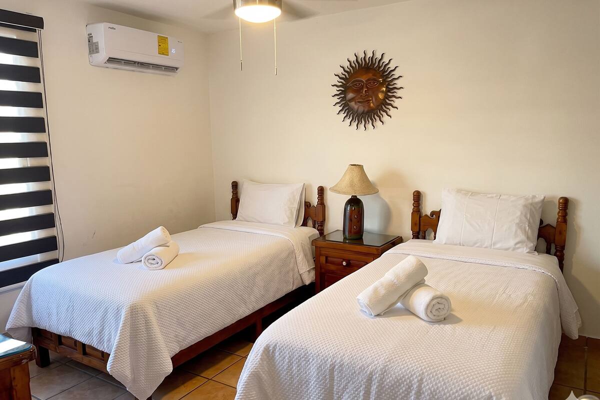 A hotel room with two beds, a nightstand with a lamp, a wall-mounted air conditioner, and a decorative sun-like object.