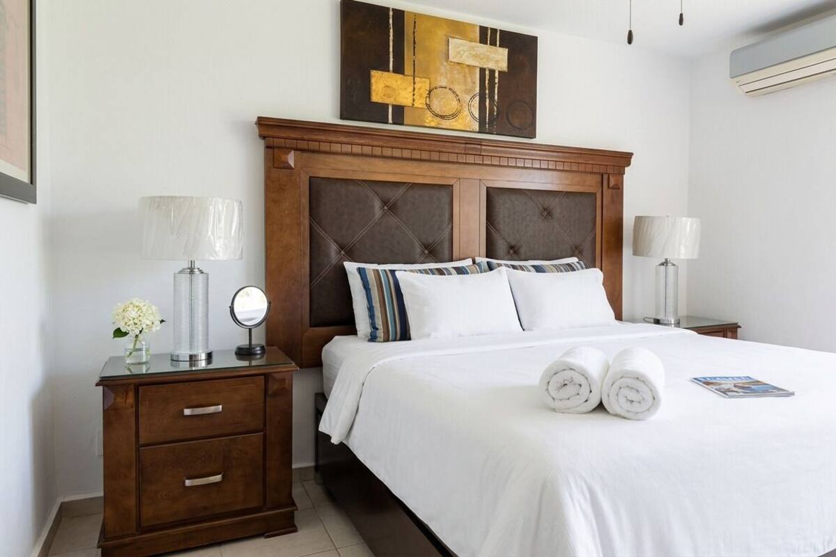 A hotel room with a large bed, wooden headboard, two bedside tables with lamps, a mirror, and a framed artwork above the bed.