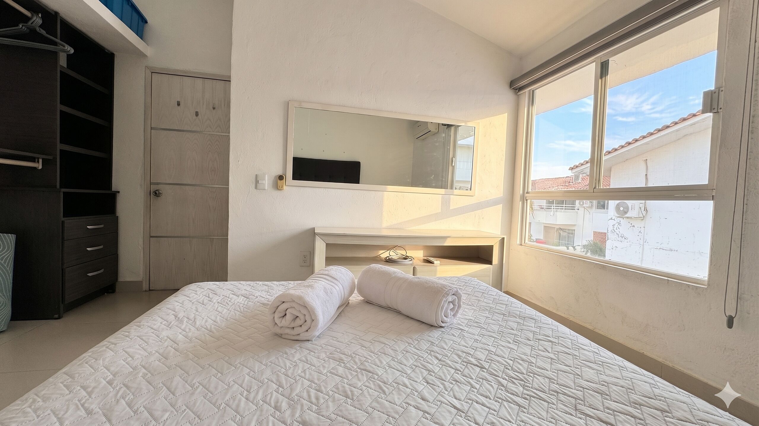 Een slaapkamer met een bed, een kaptafel, een spiegel en een raam met uitzicht op gebouwen.