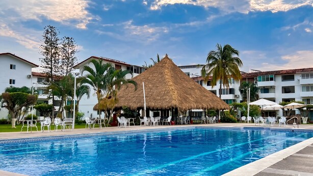 A swimming pool with white chairs and a thatched-roof structure, surrounded by buildings and palm trees. - La Marina Condominios by Monyxbnb (Puerto Vallarta)