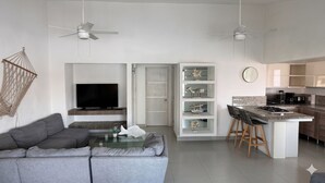 A modern living room with a gray sofa, a flat-screen TV, and a glass coffee table. There are two ceiling fans, a kitchen with a granite countertop, and a dining area with bar stools. - La Marina Condominios by Monyxbnb (Puerto Vallarta)