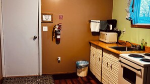 Une cuisine compacte équipée d’un four à micro-ondes, d’une cuisinière et d’un évier. Un extincteur est fixé au mur, et un panneau indique l’heure limite pour quitter les lieux.