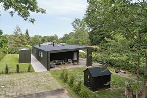 Ein modernes Haus mit überdachter Terrasse, einem kleinen Gartenhaus und einer Rasenfläche.
