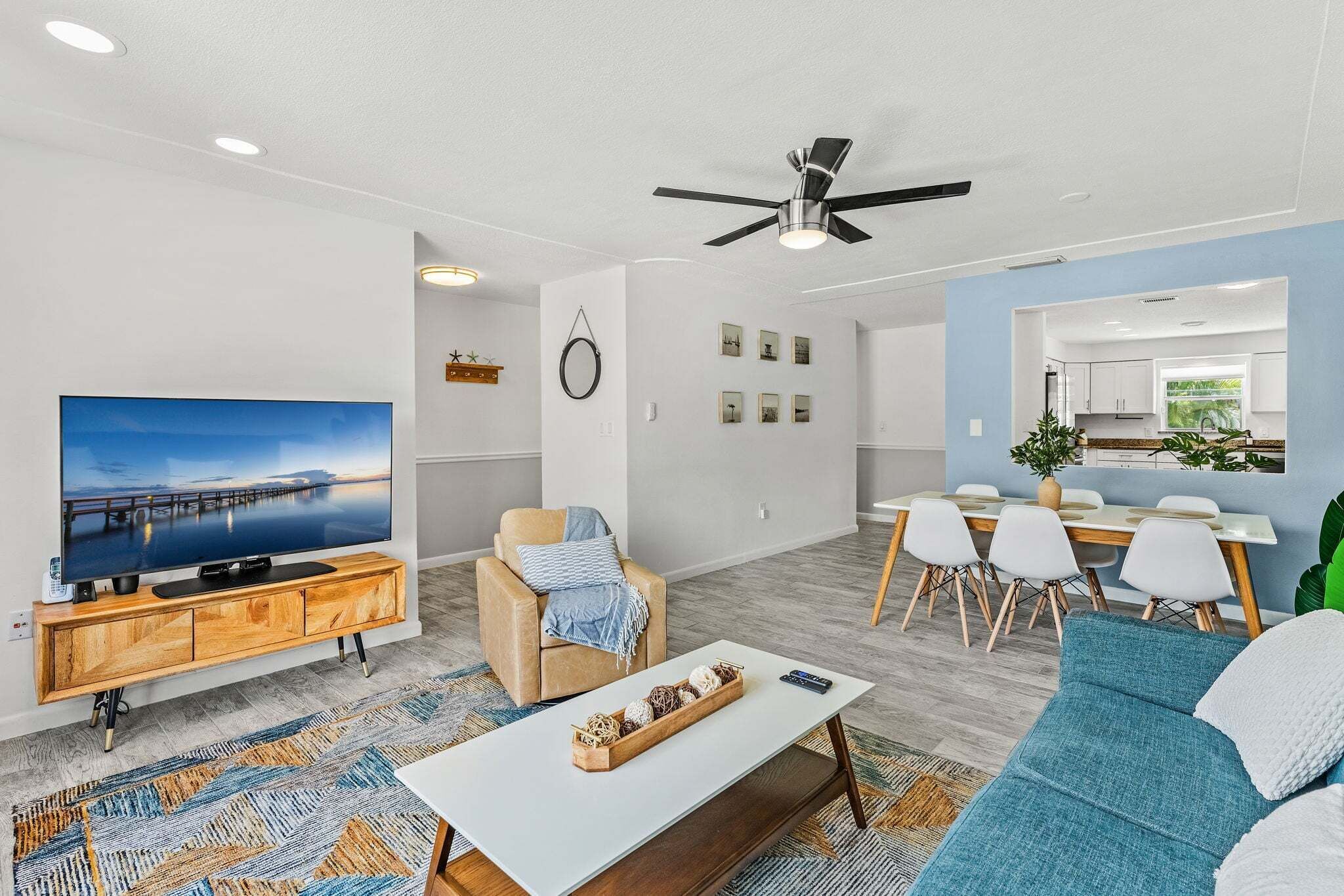 A modern living room with a flat-screen TV, a sofa, a coffee table, and a dining area with a kitchen in the background.