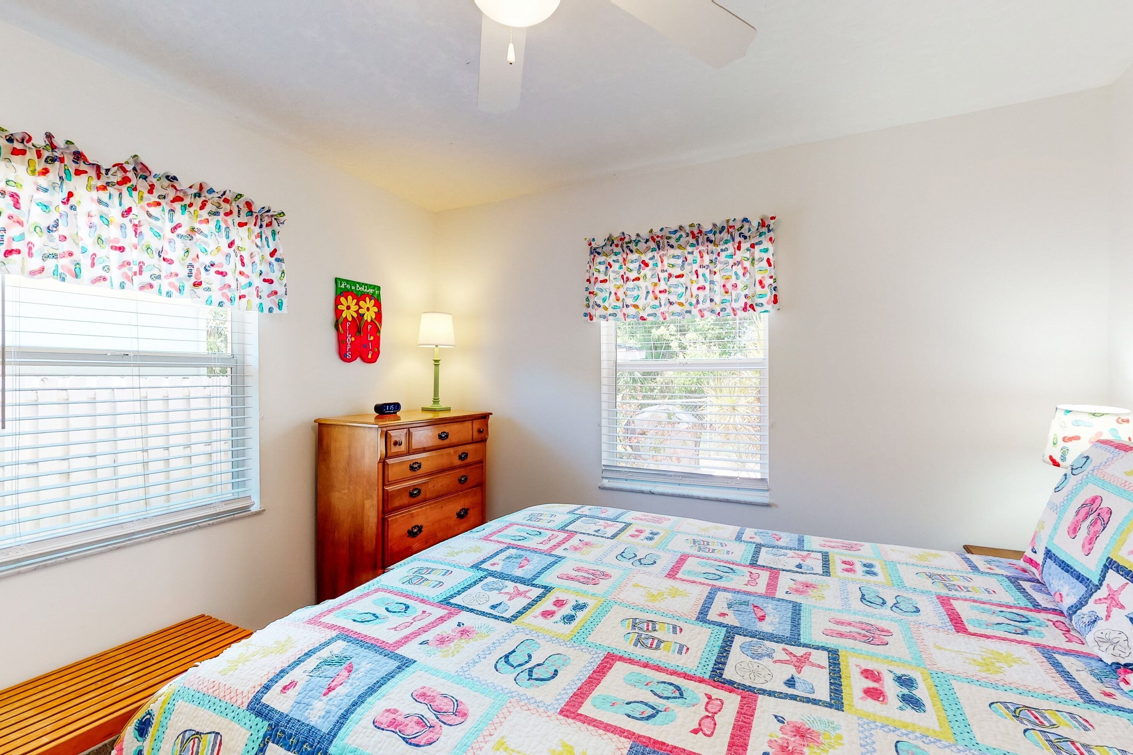 Un dormitorio con una colcha acolchada, un aparador de madera y dos ventanas con cortinas.