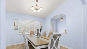 Une salle à manger avec une table en bois, des chaises blanches, un lustre et une fenêtre avec des stores.