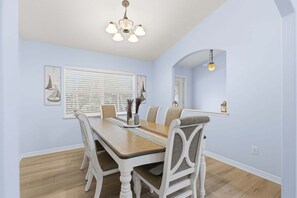 A dining room with a wooden table, white chairs, a chandelier, and a window with blinds. - 4365 9th Ln (Vero Beach)