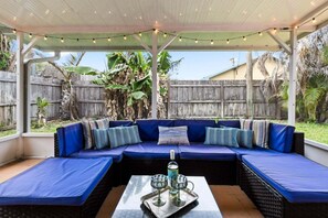 A covered outdoor seating area with a sectional sofa, a table with a tray of glasses and a bottle, and string lights. - 206 Ocean View Lane (Indialantic)