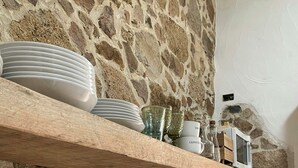 Una cocina con pared de piedra, estante de madera con platos y cristalería, pileta de acero inoxidable y horno empotrado.