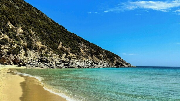Una playa con agua turquesa cristalina, orilla arenosa y un promontorio rocoso al fondo.