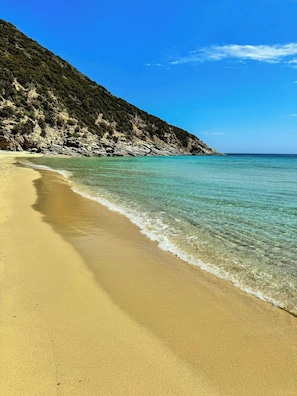 Ein Strand mit klarem türkisblauem Wasser, sandigem Ufer und einer felsigen Hügelkette im Hintergrund.