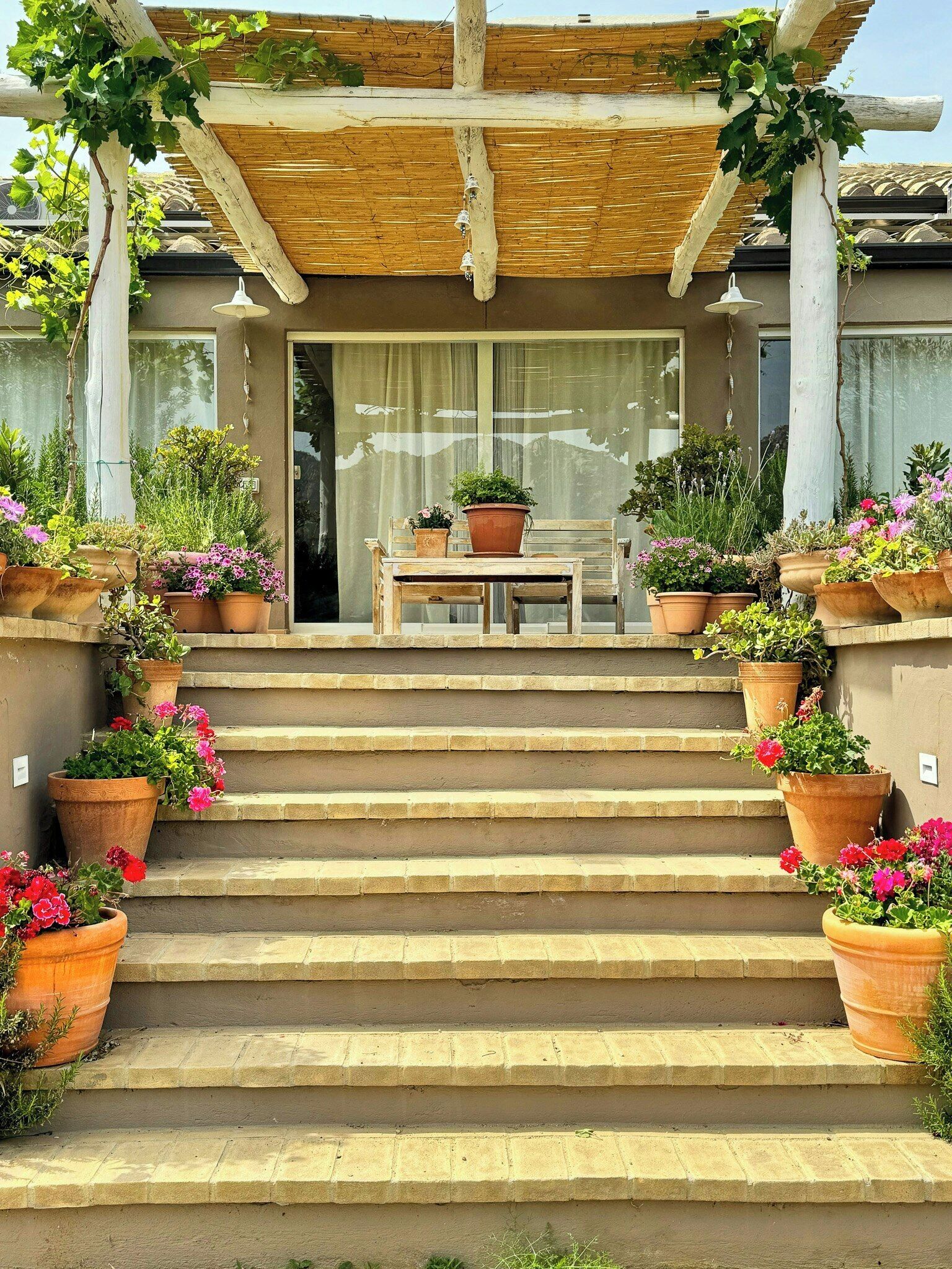 A set of stone steps leading to a wooden pergola with a table and chairs underneath, surrounded by potted plants.