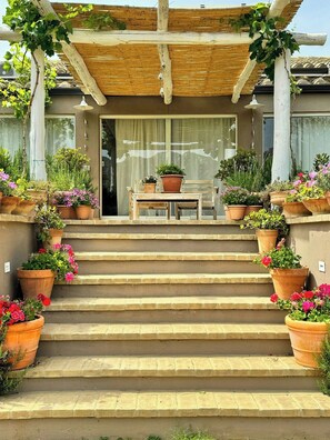 Steinstufen führen zu einer Holzpergola mit Tisch und Stühlen, umgeben von Topfpflanzen.