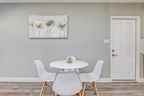 Un coin repas avec une table ronde et quatre chaises blanches, une porte blanche et un tableau accroché au mur.