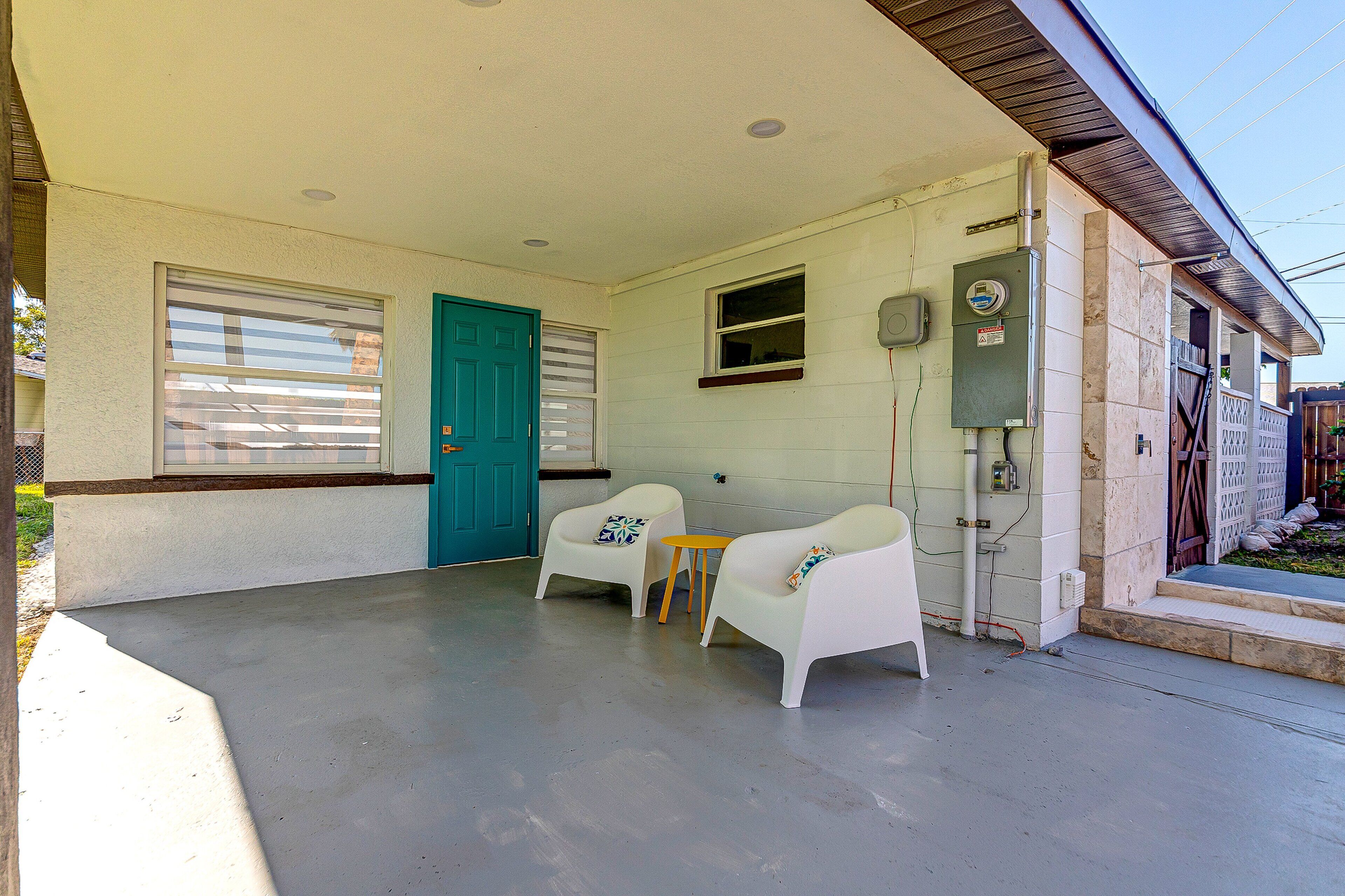 A covered porch with two white chairs and a small table, a teal door, and a window with blinds.