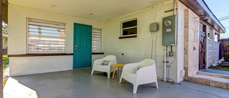A covered porch with two white chairs and a small table, a teal door, and a window with blinds.