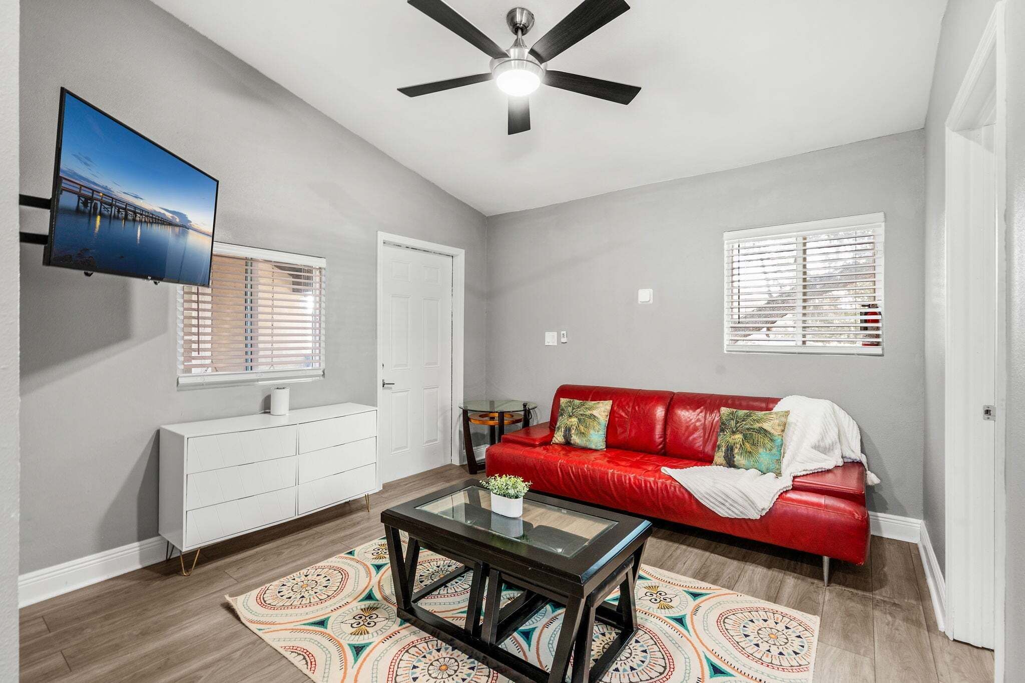 A living room with a red sofa, a glass coffee table, a TV mounted on the wall, and a ceiling fan.
