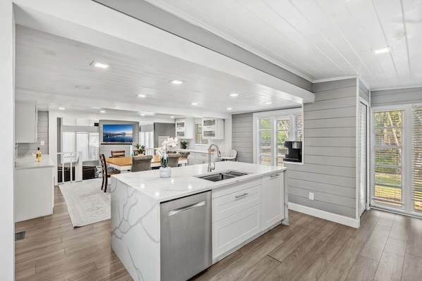 A modern kitchen with white cabinetry, a large island with a sink, and a dining area with a table and chairs. - 1055 Shady Lane (Merritt Island)