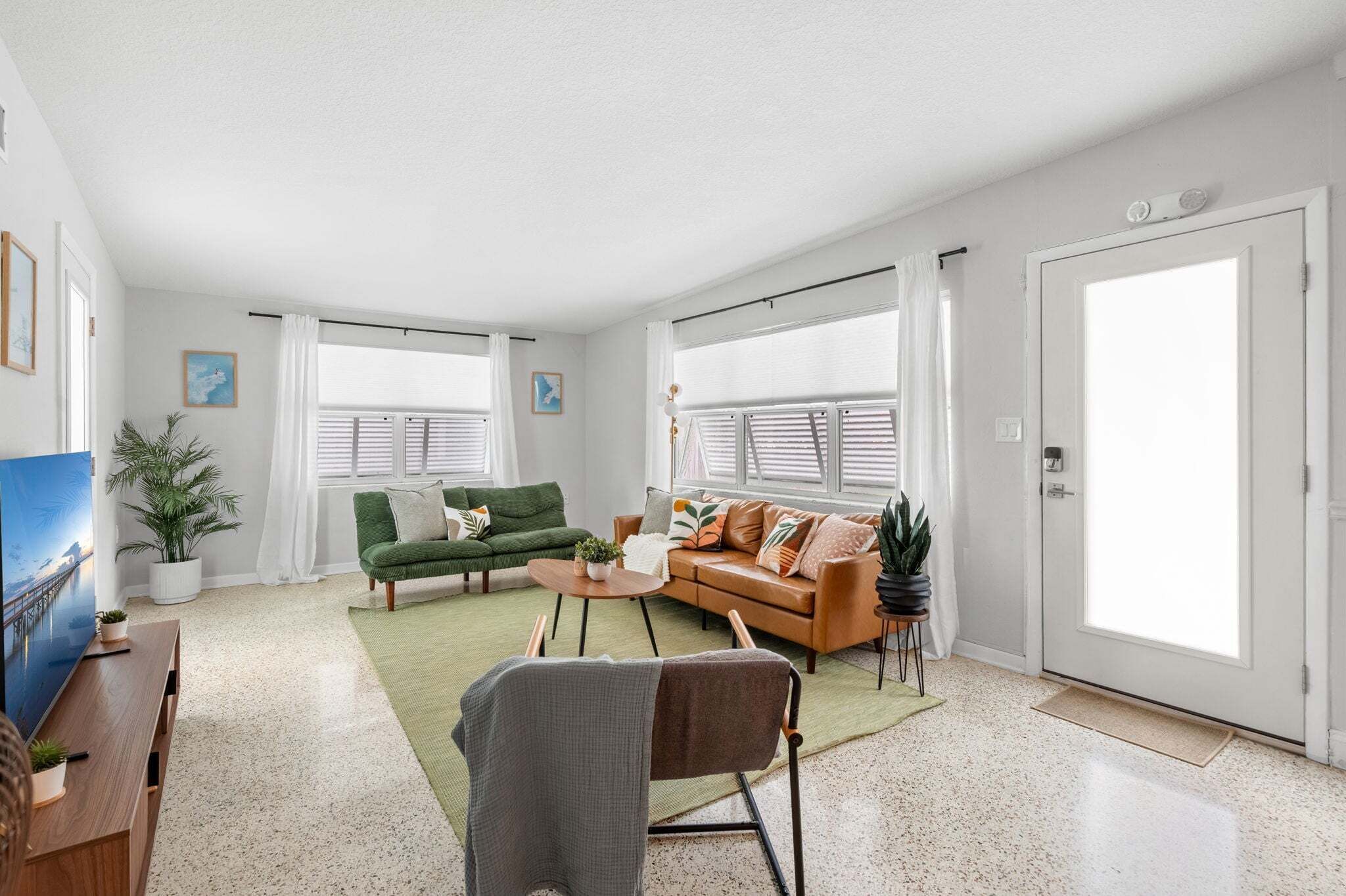 A spacious living room with a green sofa, a brown sofa, a coffee table, and a TV. There are two windows with blinds, a door, and a small table with a plant.