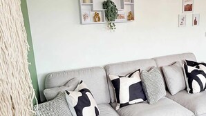 Een moderne woonkamer met een grijze bank, gekleurde kussens, een macramé wanddecoratie en een decoratieve wandplank.