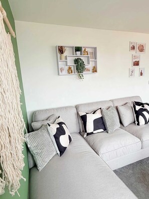 A modern living room with a grey sofa, patterned cushions, a macrame wall hanging, and a decorative wall shelf. - Penthouse with City Views (Bristol City)
