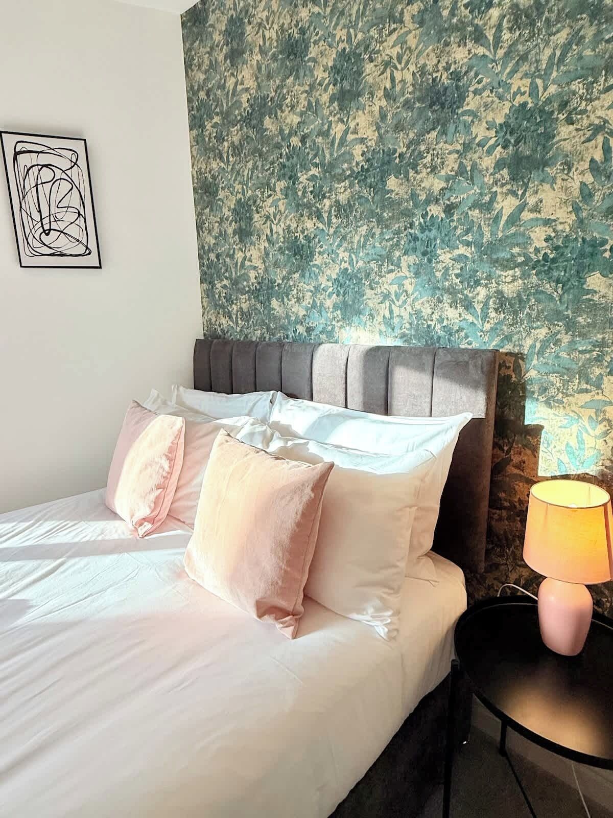 Un lit bien fait, avec des oreillers, une tête de lit et un mur orné d’un motif végétal, ainsi qu’une lampe sur une table de chevet.