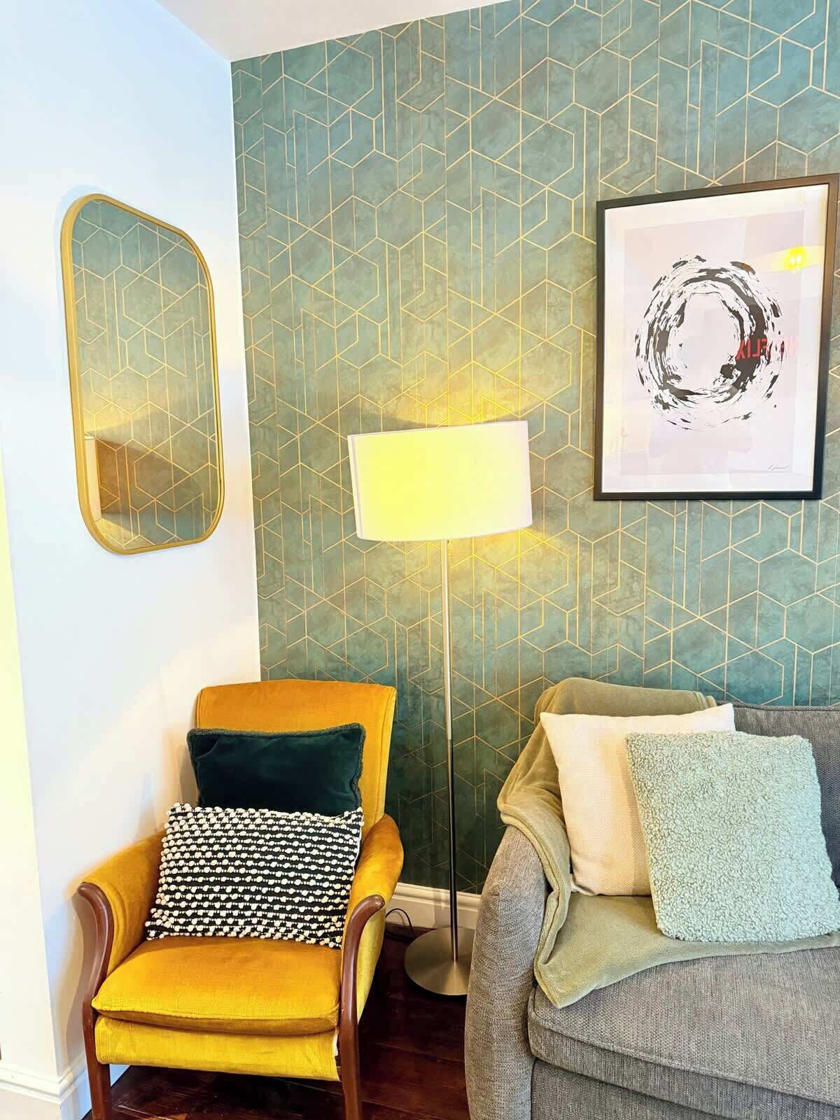 A room with a geometric patterned wall, a floor lamp, a framed abstract artwork, a yellow armchair with patterned cushions, and a grey sofa with various cushions.