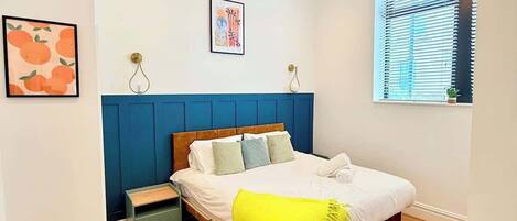 Une chambre à coucher comprenant un lit, des tables de chevet, une commode, une fenêtre avec des stores et un luminaire fixé au mur.