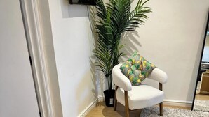 Ein Zimmer mit Holzboden, ein weißer Stuhl mit farbigem Kissen, ein brauner Ledersessel, ein Blumentopf und ein an der Wand befestigtes Fernsehgerät.