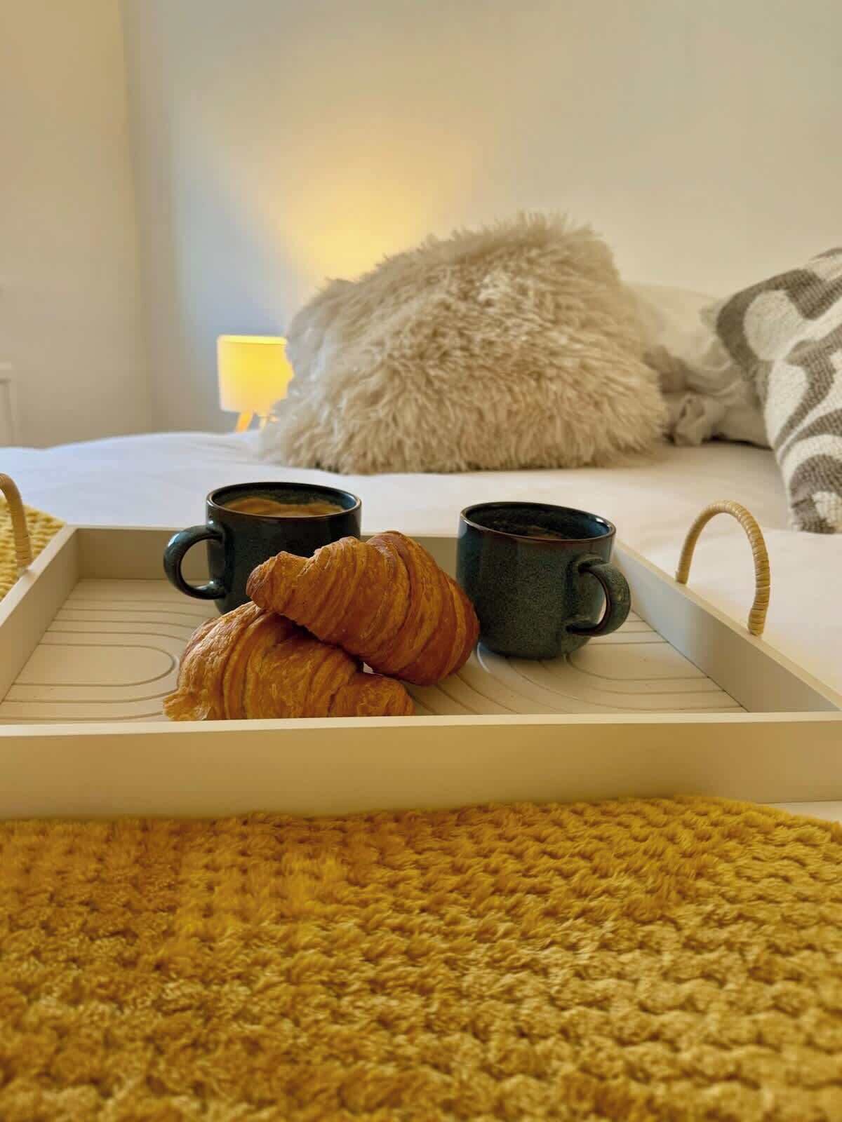 A tray with croissants and coffee cups on a bed with a fluffy pillow and a blanket.
