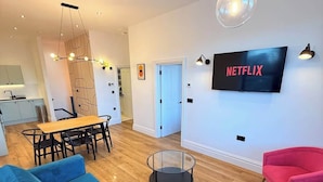 En moderne stue med en blå sofa, en mønstret lænestol, et glasbord, et spiseområde med et træbord og stole, og et vægmonteret fjernsyn der viser 'NETFLIX'.