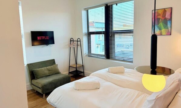 A hotel room with a bed, a sofa, a TV mounted on the wall, a window with blinds, and a painting on the wall. - Central - Free parking- Sleeps 6 - Stylish Apt (Saint Paul's)