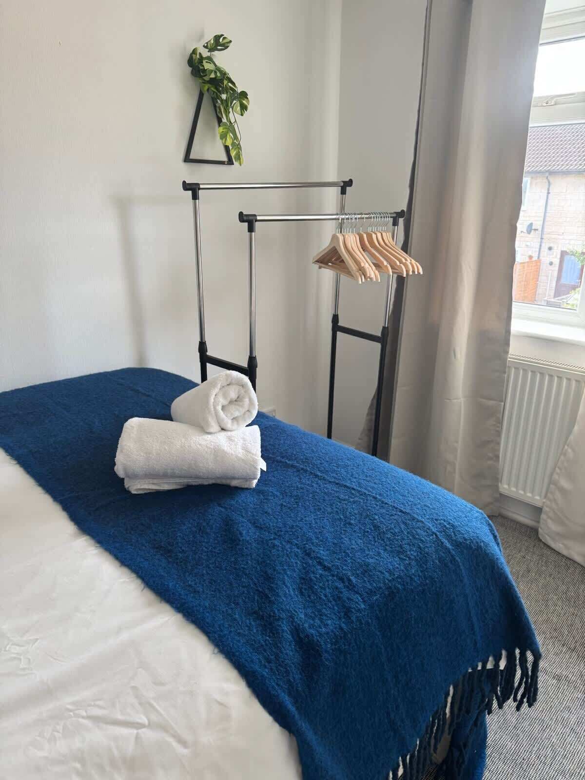 A neatly made bed with a blue blanket, a hanging clothes rack with wooden hangers, and a potted plant on the wall.