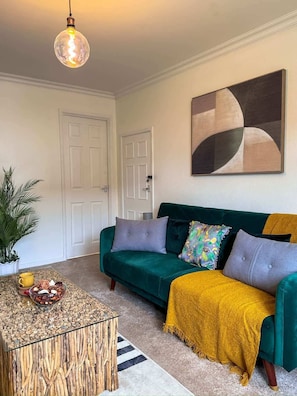 Een woonkamer met een groene bank, een gele plaid, een glazen salontafel en een hanglamp.