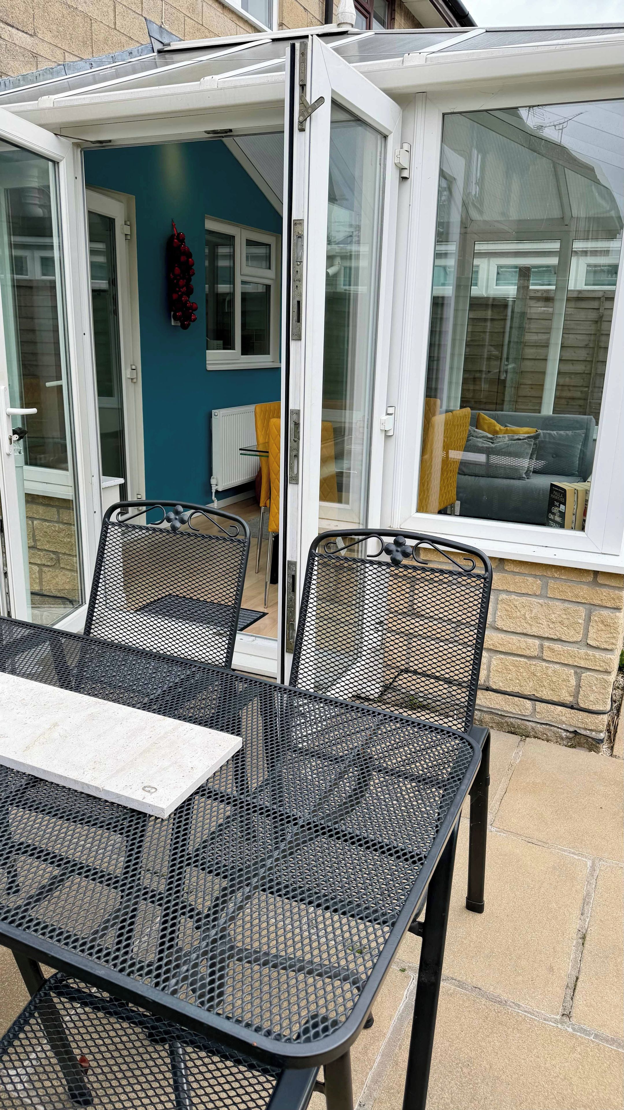 An outdoor patio with a table and chairs, a glass door leading to a room, and a view of a building through the glass.