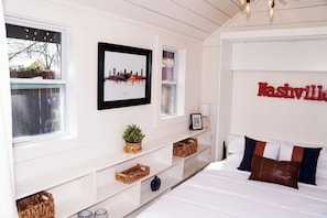 A bedroom with a bed, a cityscape picture, a wall-mounted lamp, and a shelf with baskets. - Chic Music City Retreat & Studio (Nashville)