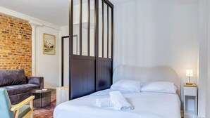 A hotel room with a bed, a seating area with a sofa and armchair, a small side table, and a brick wall. - Gathéa, studio rénové pour 2 proche du Vieux marché (Rouen)