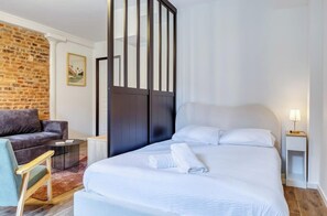 A hotel room with a bed, a seating area with a sofa and armchair, a small side table, and a brick wall. - Gathéa, studio rénové pour 2 proche du Vieux marché (Rouen)
