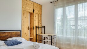 Un dormitorio con cama, armario de madera, escritorio y ventana con cortinas translúcidas.