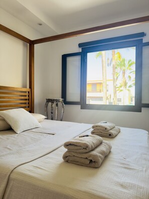 Ein Hotelzimmer mit Bett, Kissen, Tisch und Fenster mit Blick auf Palmen.
