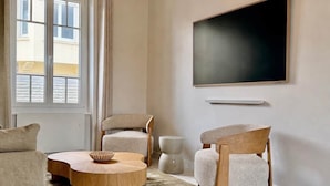 Una sala moderna con una mesa de centro redonda de madera, dos sillones tapizados, un televisor de pantalla plana montado en la pared y una ventana con persianas.