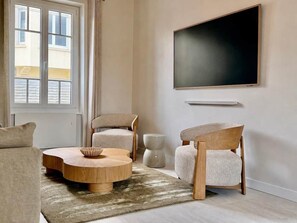 Ein modernes Wohnzimmer mit einem runden Holztisch, zwei Polsterstühlen, einem flachbildfernseher an der Wand und einem Fenster mit Jalousien.