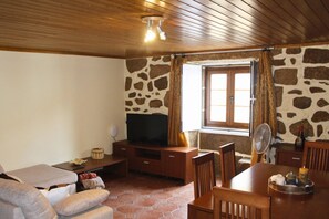 A cozy living room with a stone wall, wooden ceiling, a flat-screen TV, a dining table with chairs, and a window with curtains. - Lourença Stays – Casa Castro Sernancelhe (Sernancelhe)
