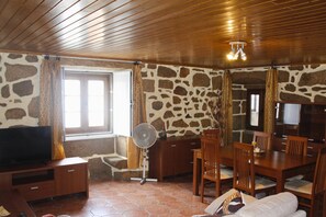 Een kamer met een stenen muur, houten plafond, een televisie, een ventilator, een eettafel met stoelen en een bank.