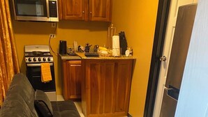 Eine kompakte Küche mit Herd, Mikrowelle und Holzschränken. Eine kleine Kücheninsel beherbergt ein Spülbecken und verschiedene Küchenutensilien. Ein Sofa befindet sich im Vordergrund.