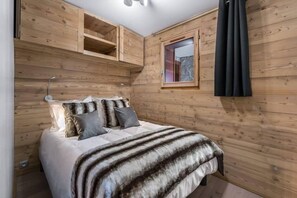 Un dormitorio con cama, almohadas, un plaid de piel sintética, paredes de madera, una ventana y una cortina.