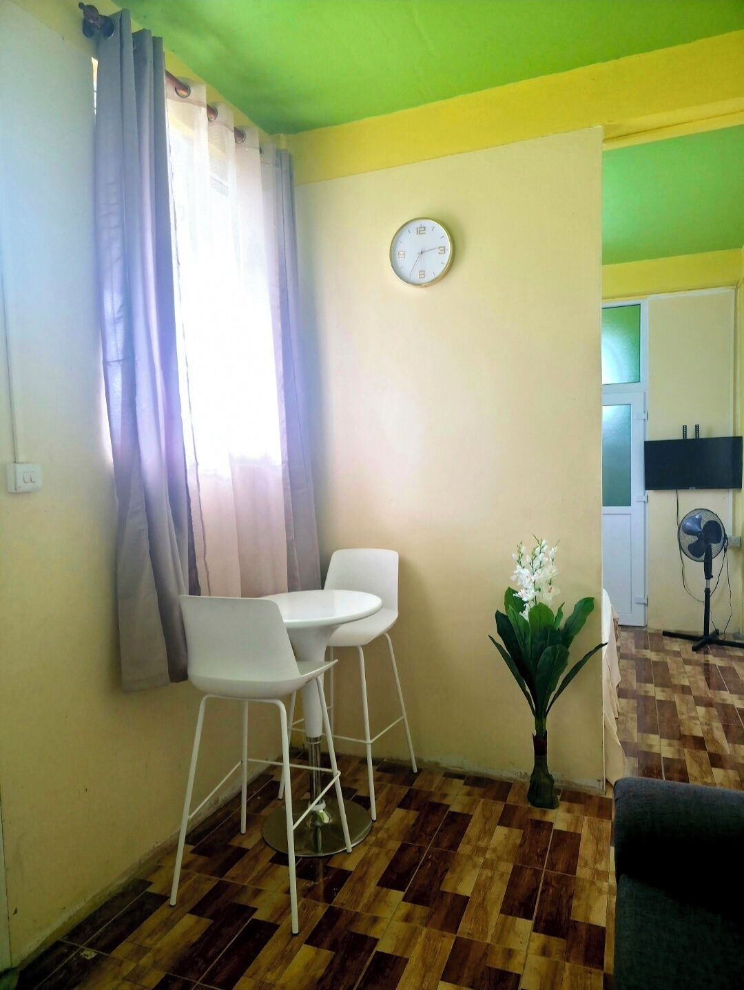 A room with a dining table and chairs, a clock on the wall, a potted plant, and a fan.